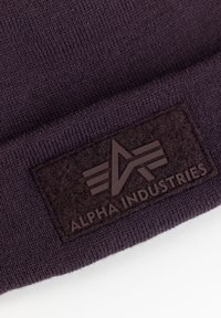 Dark purple knit beanie featuring a textured black felt patch with embossed logo reading "ALPHA INDUSTRIES" and geometric design elements.