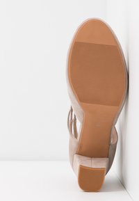Chaussure beige en daim à talon avec une semelle en caoutchouc marron appuyée contre un mur blanc, taille 37, montrant le détail de la bande de roulement.