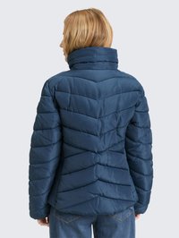 Blond persoon die een marineblauwe doorgestikte donsjas met hoge kraag draagt, staand met de rug naar voren, gecombineerd met blauwe spijkerbroeken.