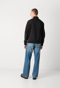 Black lightweight bomber jacket with a standing collar, elastic cuffs, and waistband, paired with loose-fitting blue denim jeans.