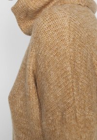 Maglione a collo alto in maglia chunky di colore beige, con una texture morbida e una vestibilità ampia. La vista laterale mette in evidenza il colletto a costine e il dettaglio del motivo in maglia.
