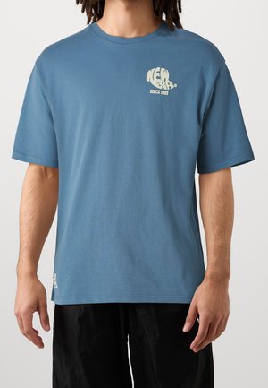 Homme portant un t-shirt bleu à manches courtes avec le texte "New Era Since 1920" en crème sur la partie supérieure gauche de la poitrine, associé à un short noir.