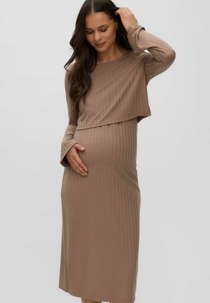 Femme enceinte aux longs cheveux foncés portant une robe marron clair côtelée à manches longues, touchant délicatement son ventre et ses cheveux.