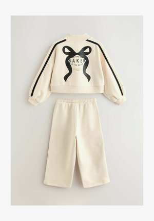 Krém színű rövidített pulóver fekete szalagmintával és felirattal, mellékelve lazán szabott nadrággal, amelynek derékrésze elasztikus.