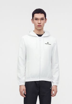 Un mannequin masculin se tient devant un fond uni, vêtu d’une veste blanche à capuche et à fermeture éclair et d’un pantalon noir.