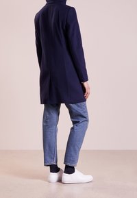 Navy wool coat with a high collar, tailored fit, single vent at the back, paired with light blue jeans and white sneakers.