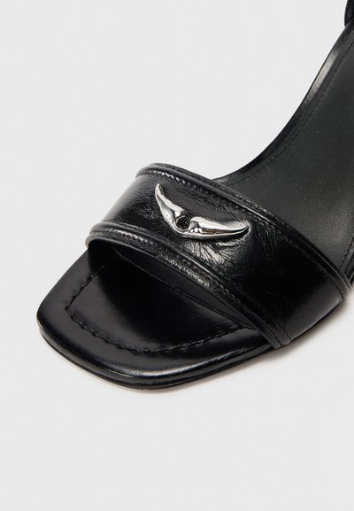 Čierna sandála s otvorenou špičkou, hranatým tvarom špičky, lesklým koženým zvrškom, dvojitými remienkami a strieborným krídlovým ozdobným prvkom vpredu.