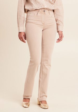 Person trägt hellbeige, eng anliegende Hose, passend gestreiftes Hemd, in die Hose gesteckt, und goldene Blockabsatzschuhe, steht vor neutralem Hintergrund.