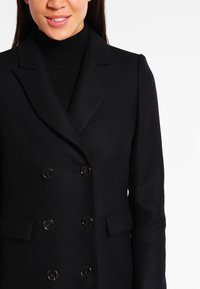 Sort dubbelbrystet frakke med klippelapper, struktureret stof og blanke knapper. Har sidelommer og en tætsiddende silhuet.