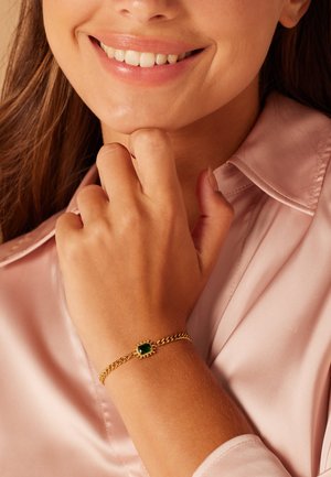 Gold bracelet featuring a rectangular green stone, braided chain design, worn on a hand against a pink satin fabric.