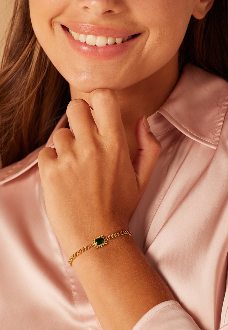Bracelet en or avec une pierre verte rectangulaire, design en chaîne tressée, porté sur une main sur un tissu en satin rose.