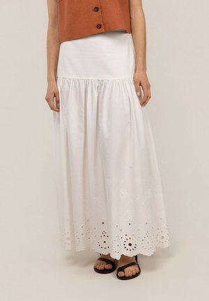 Mujer con una falda larga blanca con detalles de ojales y recortes florales y sandalias planas negras, combinada con un top marrón sin mangas.