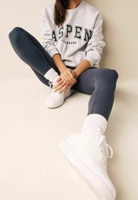 Felpa grigia con il testo "ASPEN", abbinata a leggings scuri e sneakers alte bianche. Il modello è seduto con le gambe incrociate e indossa calzini bianchi.