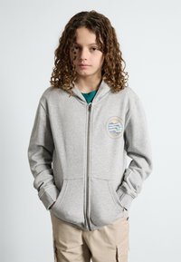 Moleton cinza com fecho, feito de uma mistura de algodão, apresentando um bolso frontal e um logotipo circular no lado esquerdo do peito. Mangas longas com punhos canelados.