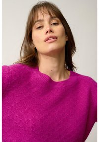Pull en tricot fuchsia avec un col rond, présentant une surface texturée et des poignets côtelés. Tissu doux avec une coupe ample.