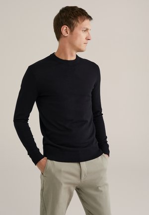 Man i en svart långärmad tröja och beige byxor, står med händerna i fickorna och tittar åt sidan mot en neutral bakgrund.