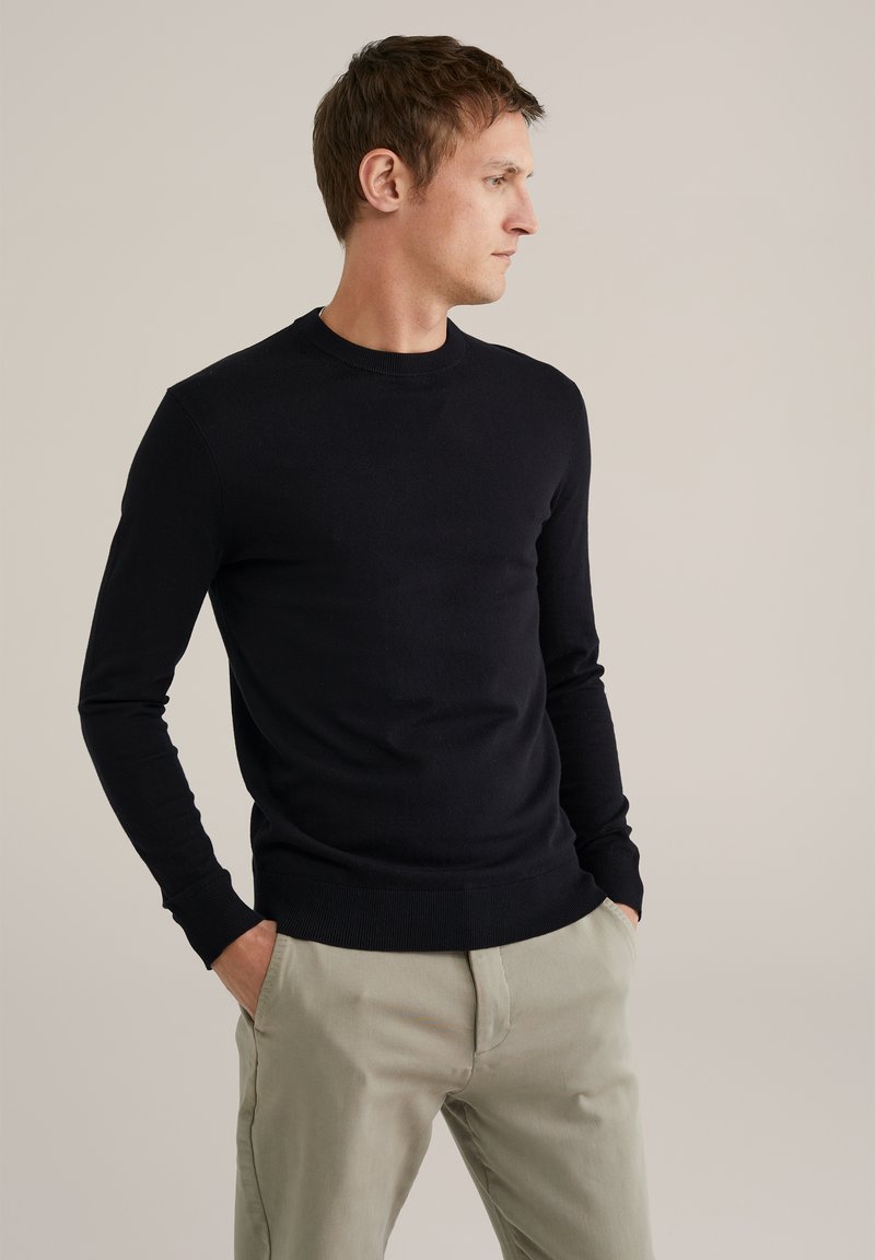 Hombre que lleva un suéter negro de manga larga y pantalones beige, de pie con las manos en los bolsillos, mirando hacia un lado contra un fondo liso.
