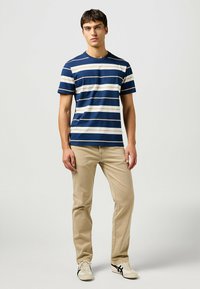 T-shirt a righe in navy, bianco e beige con maniche corte, abbinata a pantaloni beige chiaro. Vestibilità standard, design a girocollo, materiale in cotone.