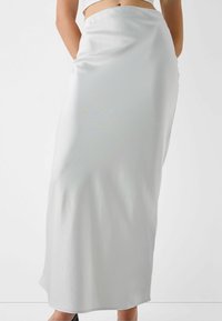 Bílá saténová maxi sukně s hladkým povrchem, rovného střihu a asymetrickým lemem, doplněná černými podpatky.