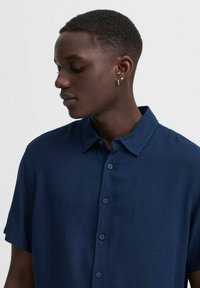 Námořnická košile s krátkými rukávy na knoflíky vyrobená z lehkého materiálu; má standardní límec a pět knoflíků vpředu.