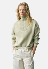 Light green sweater with a high collar and ribbed pattern, paired with cream-colored, relaxed-fit pants. Soft texture and loose silhouette.