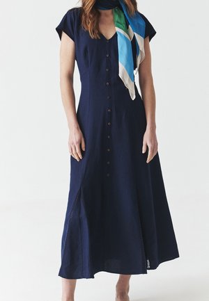 Femme portant une robe bleu marine boutonnée sur le devant avec des manches courtes et une écharpe bleue, verte et blanche autour du cou.