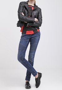 Chaqueta de motorista de cuero negro con hombros acanalados, llevada sobre una blusa roja y negra. Jeans de mezclilla azul con un diseño ajustado y detalles en panel. Zapatos negros.