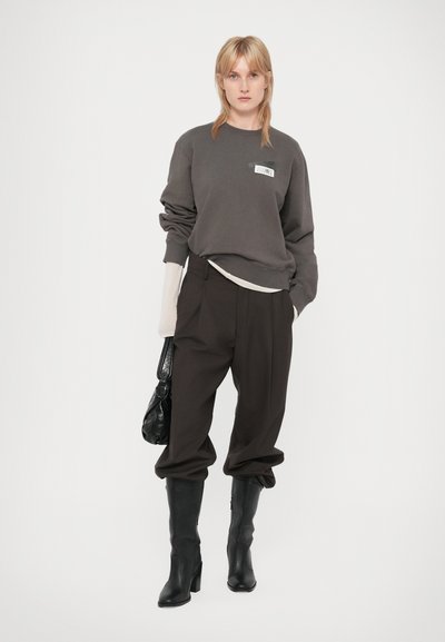 Mujer joven con sudadera gris oscuro, pantalones oscuros holgados metidos en botas de tacón negro, sosteniendo un bolso negro, de pie contra un fondo blanco.