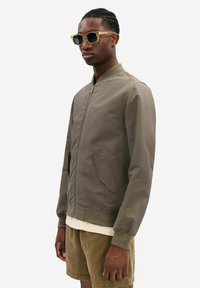 Olive green bomber jacket with a zip front, ribbed cuffs, and a rounded collar; features two side pockets and a smooth, matte texture.