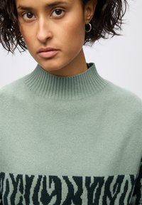 Ljusgrön ulltröja med en ribbad hög krage och svart djurmönster i nederkant. Mjuk textur, figurnära design.