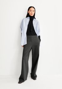 Helles blaues Hemd mit Knopfleiste, über einem schwarzen Rollkragenpullover getragen, kombiniert mit weit geschnittenen, dunkelgrauen Hosen und schwarzen Schuhen mit Nietenakzenten.