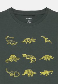 Dunkelgrünes Baumwoll-T-Shirt mit gelben Dinosaurierillustrationen, darunter ein T-Rex, Triceratops und Pterodaktylus, in verschiedenen Positionen.