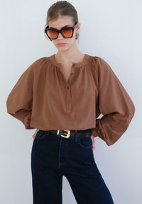 Žena oblečená v nadměrné hnědé košili na knoflíky, tmavě modrých džínách s vysokým pasem, černém opasku a velkých hranatých slunečních brýlích, stojící s rukama na bocích.