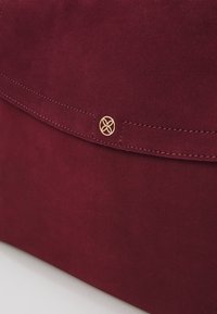 Rabattre de sac à main en daim rouge foncé avec coutures visibles et petit emblème décoratif rond en or représentant un motif à quatre pétales.