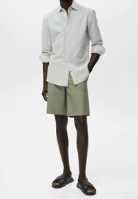 Camicia a righe con bottoni in azzurro chiaro e bianco, abbinata a pantaloncini verde oliva. Il modello indossa sandali neri, mostrando un abbigliamento estivo casual.