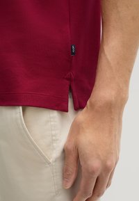 Burgunder T-Shirt mit seitlichen Schlitzen, aus weicher Baumwolle. Etikett sichtbar an der linken Seitennaht. Kombiniert mit hellbeigen Hosen.