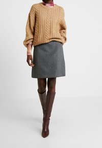 Henkilö, jolla on päällään beige palmikkoneule, harmaa polvipitkä hame, maroonit polvipituiset saappaat ja vaaleanpunainen sidontakuvioinen poolopaita neuleen alla.