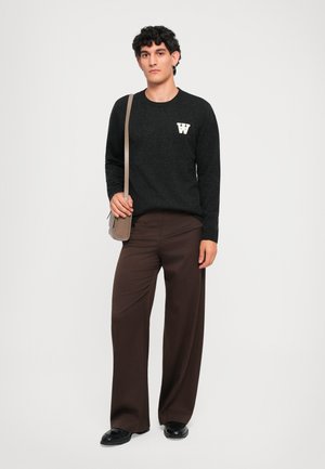 Black knit sweater with white "W" logo on the left chest; paired with loose brown trousers and a small beige crossbody bag.