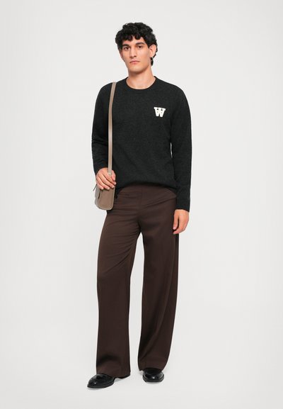 Schwarzer Strickpullover mit weißem "W"-Logo auf der linken Brust; kombiniert mit lockeren braunen Hosen und einer kleinen beigen Umhängetasche.