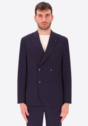 Uomo che indossa un blazer doppiopetto blu navy scuro sopra una camicia bianca su uno sfondo chiaro e uniforme.