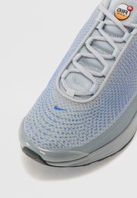 Nike Air Max sneaker in light gray mesh with blue accents, rounded toe, flat laces, and a glossy gray rubber sole. Features "Air Max" logo.