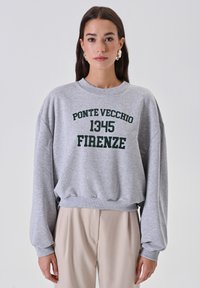 Donna che indossa un felpa grigia con la scritta "Ponte Vecchio 1345 Firenze" e pantaloni beige, in piedi contro uno sfondo chiaro uniforme.
