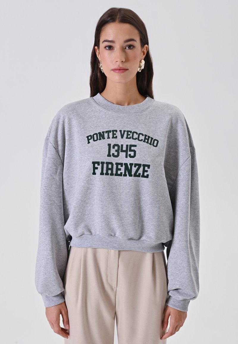 Donna che indossa un felpa grigia con la scritta "Ponte Vecchio 1345 Firenze" e pantaloni beige, in piedi contro uno sfondo chiaro uniforme.