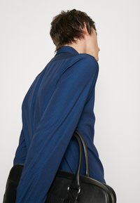 Marinblå skjorta med en strukturerad mönster, lång ärm och en figurnära design, tillsammans med en svart läderväska som hålls i handen.