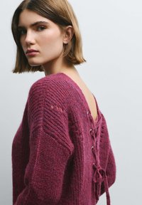 Jersey de punto en color burdeos intenso con mangas acanaladas texturizadas y detalle de cordones en la parte trasera. Presenta una espalda abierta con ojales y lazos.