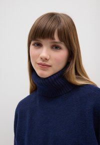 Maglione a collo alto di un blu scuro, con un texture morbida, caratterizzato da una vestibilità comoda e maniche lunghe. Lo sfondo è bianco semplice.