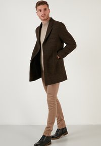 Abrigo de cuadros marrón con cuello, diseño de doble botonadura y dos bolsillos laterales. Un par de pantalones slim-fit en tono caqui y botas negras completan el look.