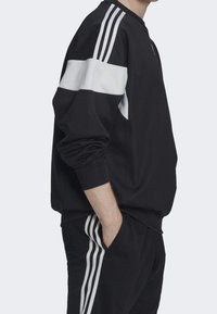 Svart sweatshirt med vita horisontella ränder på axlarna, tillverkad av ett slätt material med ribbade manschetter och en avslappnad passform.