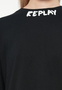 Must musta, mis on valmistatud puuvillast, ümar kaelus ja valge kiri "REPLAY" ülemisel seljal. Sile tekstuur.
