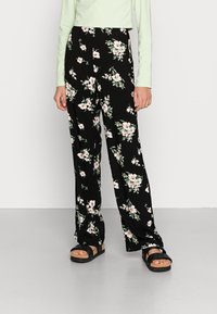 Mustat kukkahousut, joissa on väljähkö istuvuus ja valkoisia sekä vaaleanpunaisia kukkakuvioita. Yhdistetty vaaleanvihreään pitkähihaiseen yläosaan ja mustiin korollisiin sandaaleihin.